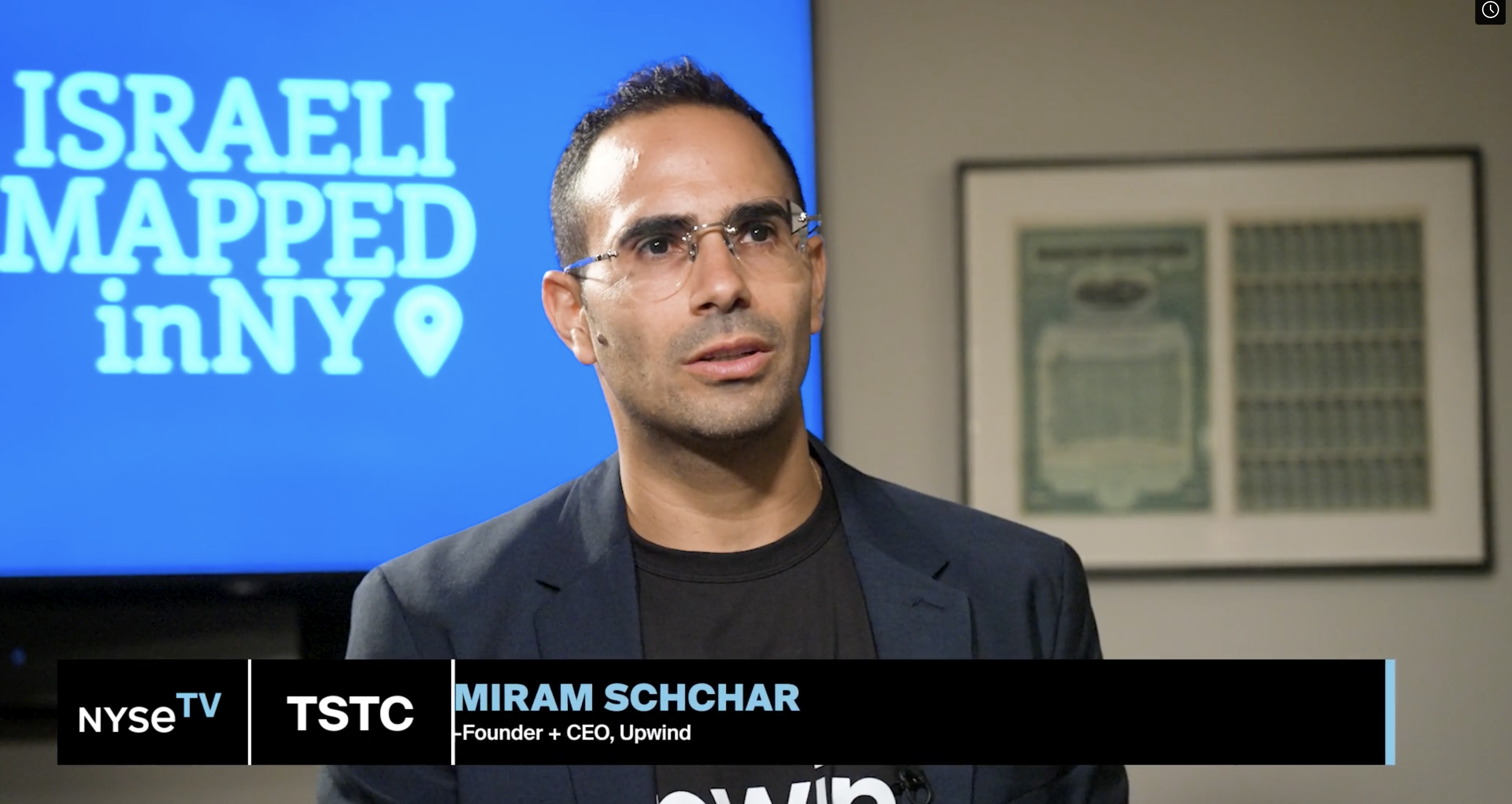 A person wearing glasses and a black shirt is speaking in front of a screen displaying the text Israeli Mapped in NY. A text banner reads, NYSETV TSTC Miram Schchar, Founder + CEO, Upwind. A framed document is on the wall in the background.